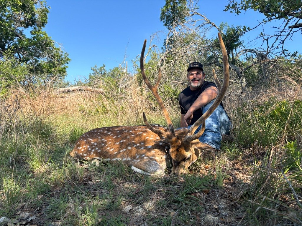 Hunter wearing LWE cap sitting beside trophy spotted Axis Deer buck in live oaks