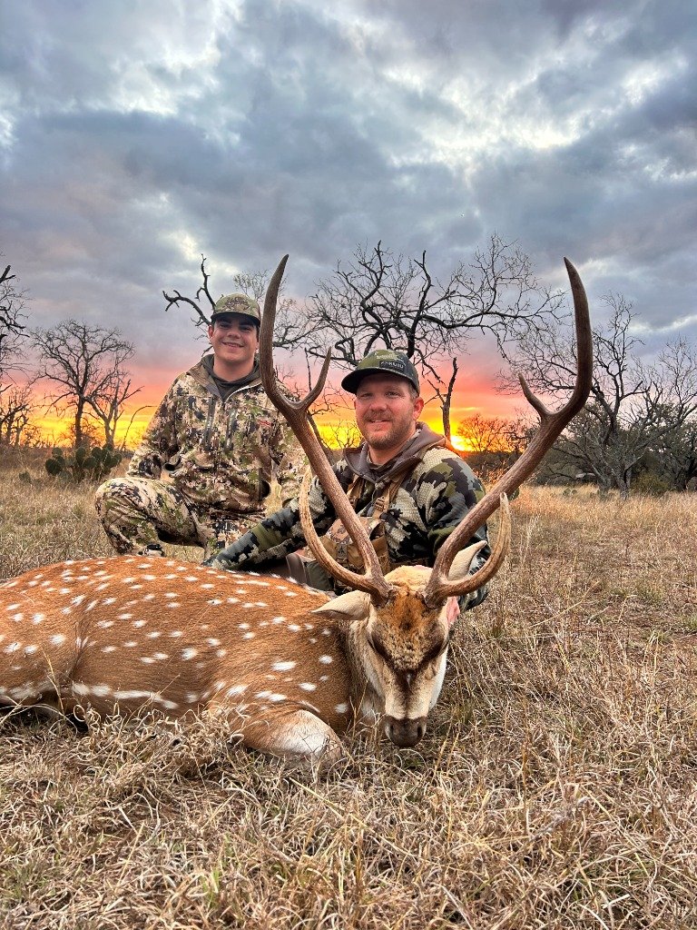 Two hunters smiling behind trophy spotted Axis stag beneath fiery orange sunset