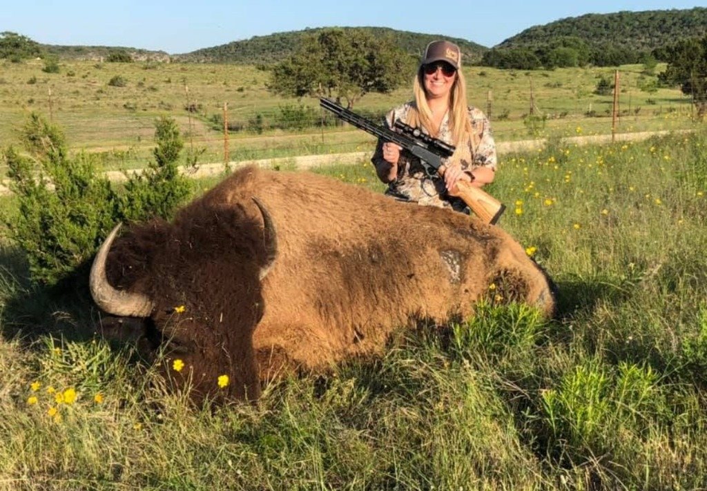 Female hunter holding lever-action rifle standing beside massive Bison bull in yellow wildflower pasture