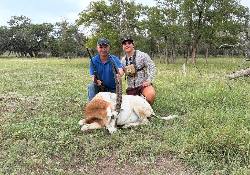 Two hunters smiling beside trophy Scimitar-horned Oryx in Texas live oak pasture